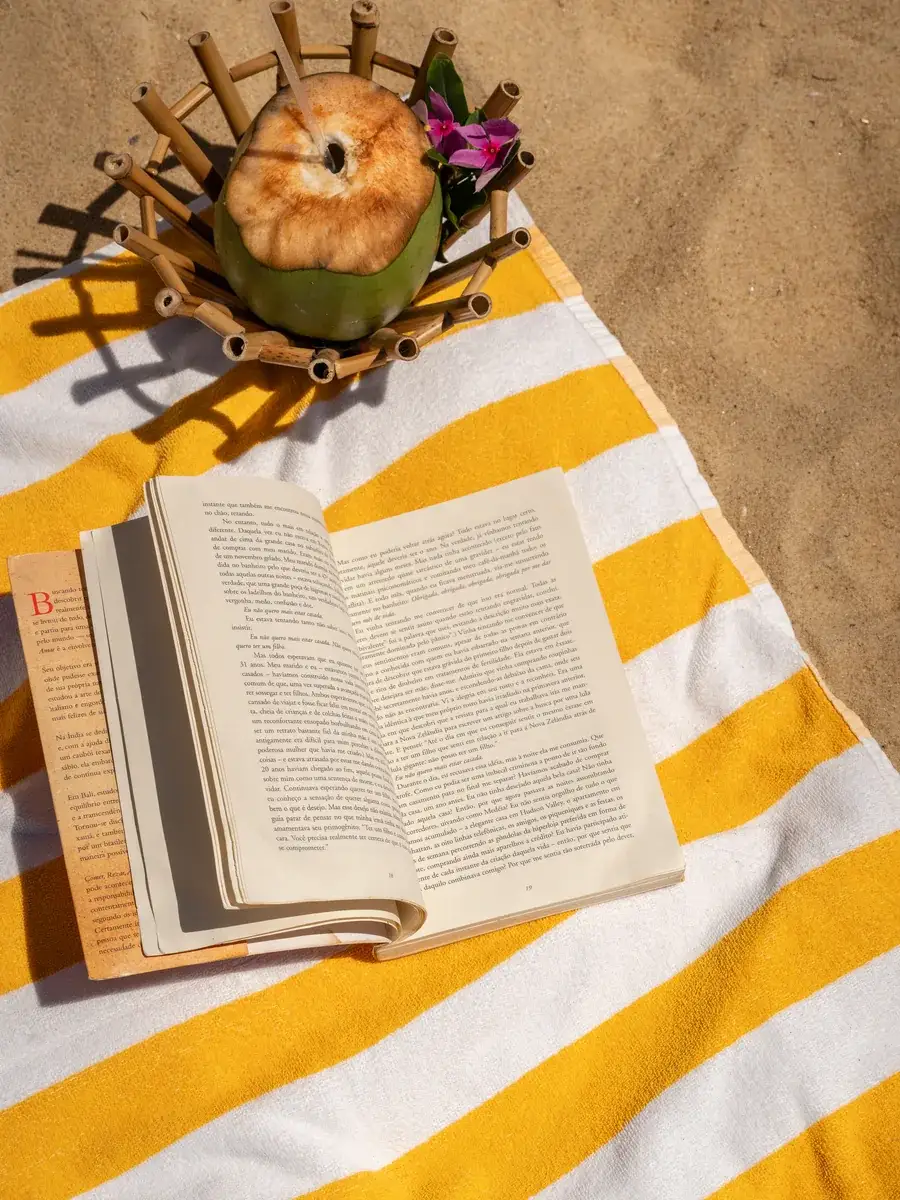 Livro aberto sobre uma canga listrada de amarelo na areia, ao lado de um coco numa cesta de palha