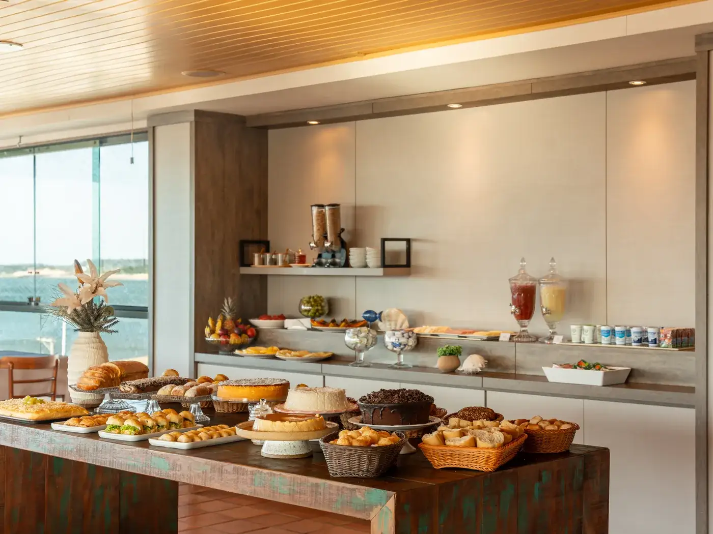 Buffet do café da manhã montado, com bolos, pães e frutas, e a janela para o mar à esquerda