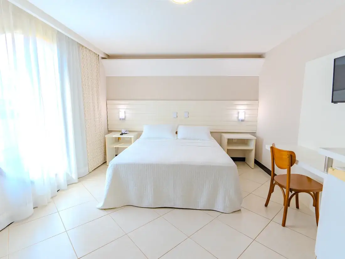 Quarto duplo com cama queen, enxoval branco, cadeira de madeira e TV