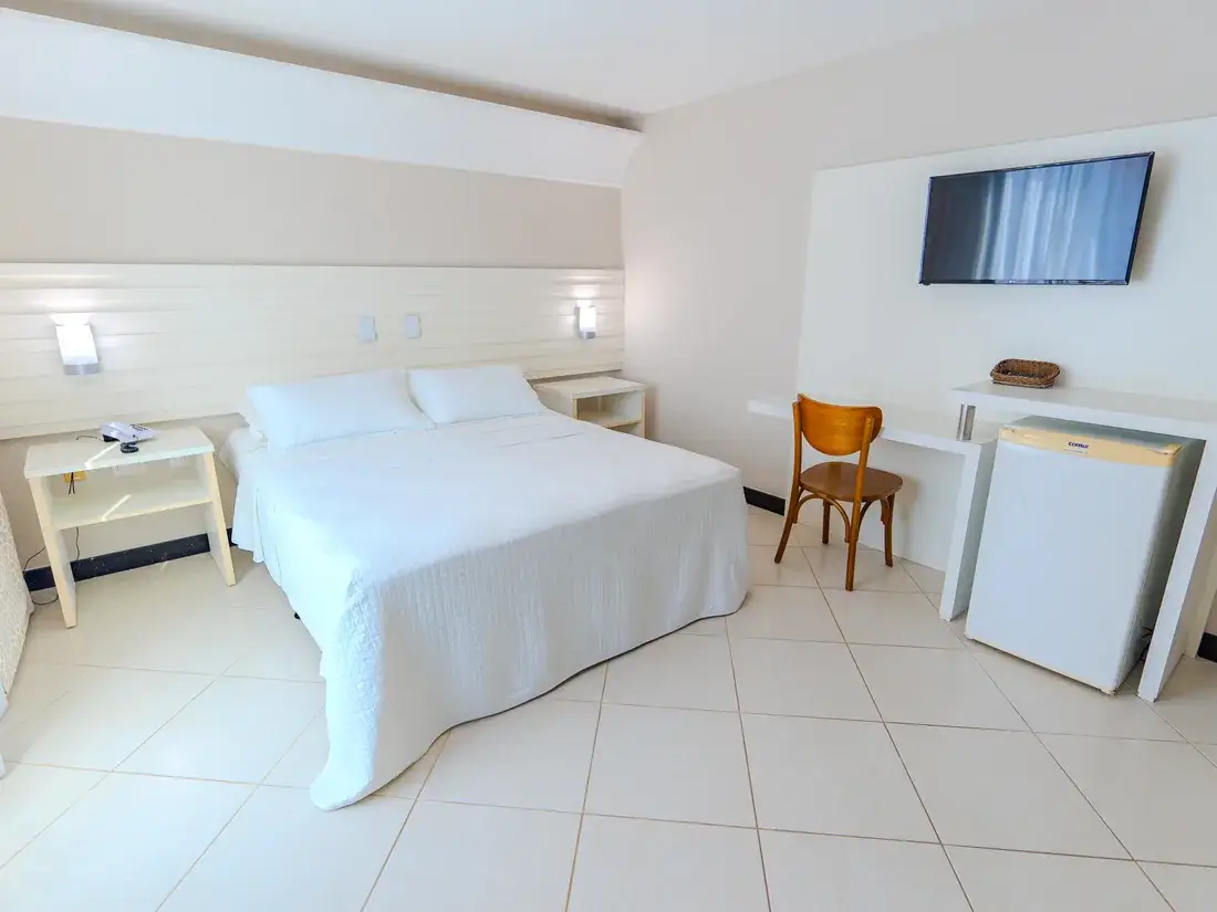 Cama queen com as luminárias acesas, a escrivaninha com cadeira de madeira, o frigobar e a TV na parede