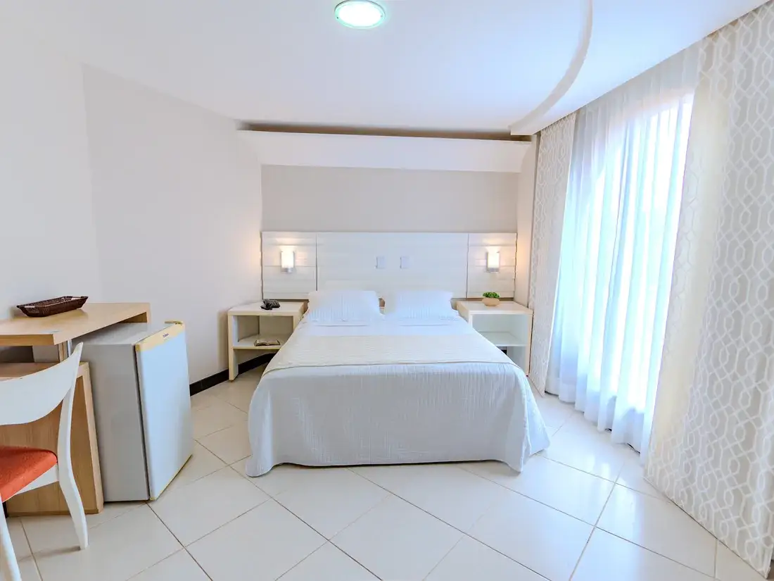 Quarto duplo em plano aberto, com escrivaninha, frigobar e a cama ao fundo