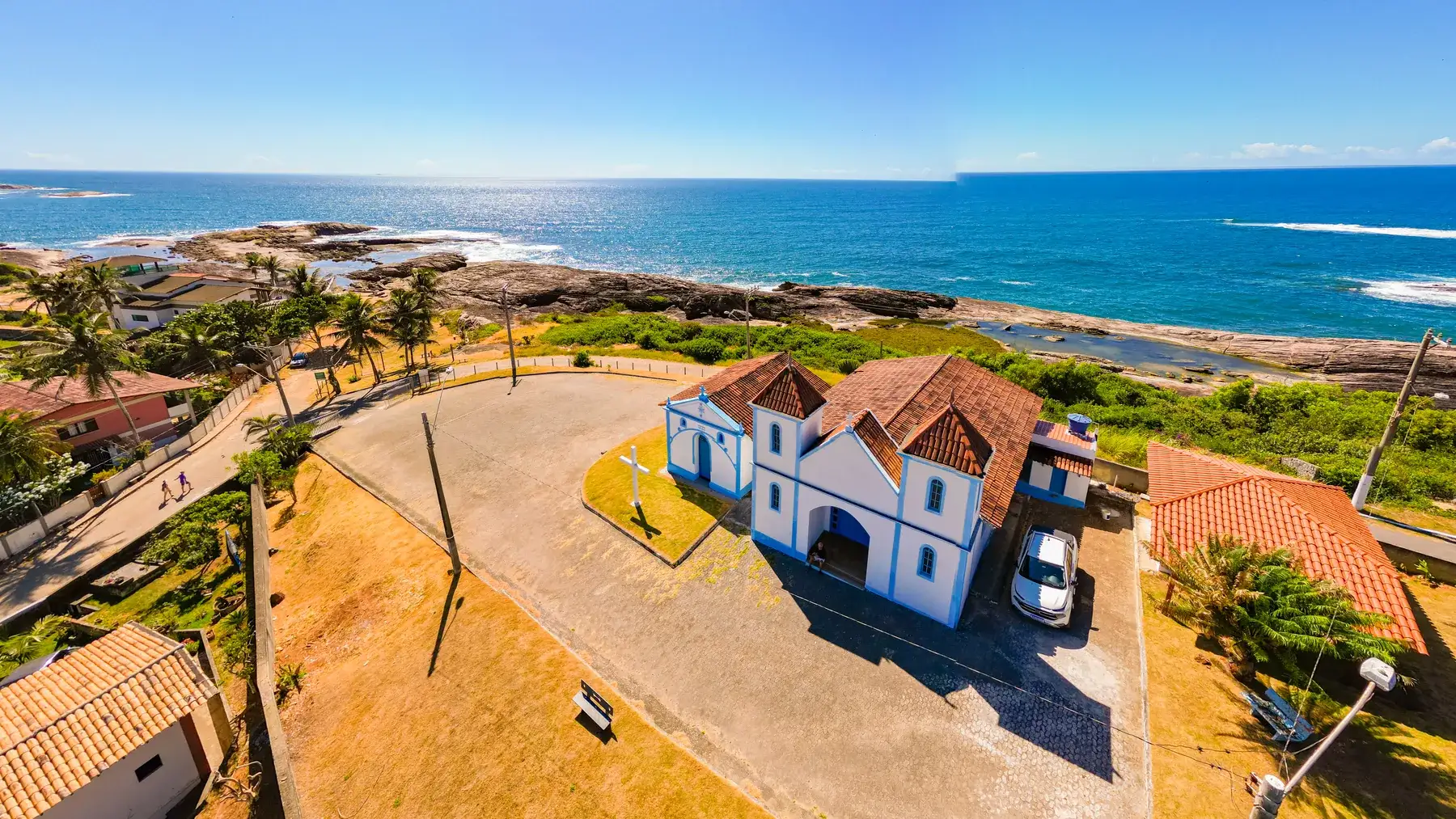 A Igreja de Sant'Ana vista do alto, branca e azul com telhado de barro, no fim da orla com o mar aberto atrás