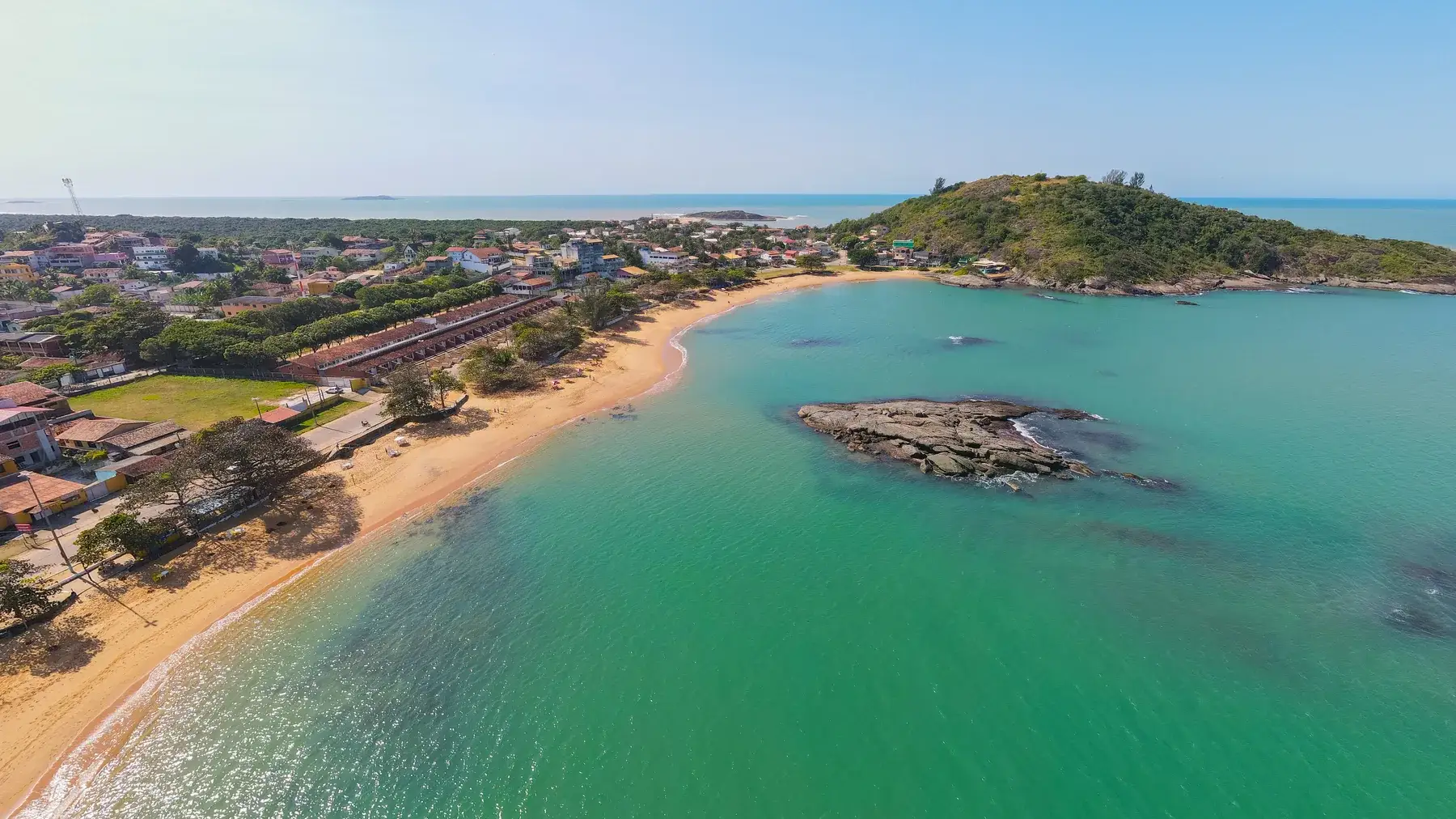 Praia de Setiba vista de drone, com a água verde e parada, uma ilhota de pedra e a faixa de areia com árvores